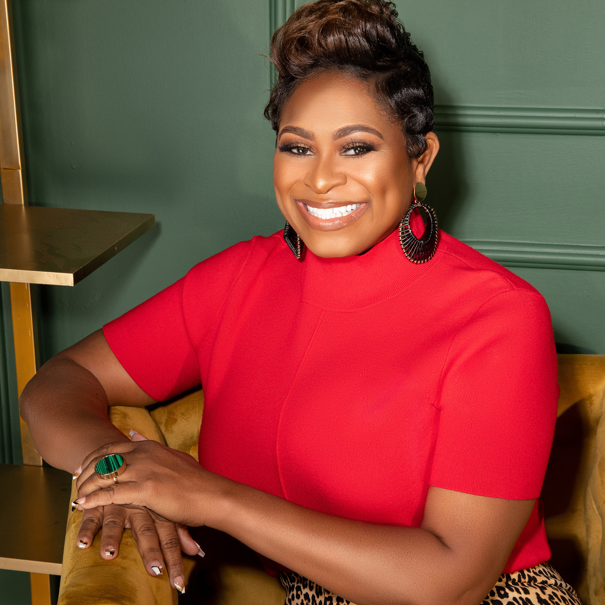 Charell G. Coleman sitting in a mustard yellow chair in a green room with a gold bookshelf to her left. She is wearing a red short sleeve top, leopard print skirt and black hoop earrings. She is leaning on the right arm rest with her left hand resting on her right while smiling at the camera.