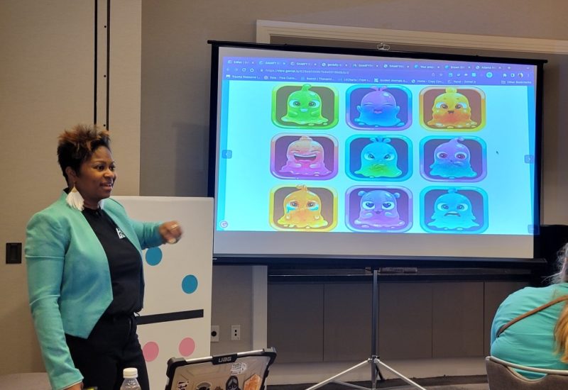 Charell G. Coleman standing at the front of a classroom to the right of a projector screen, facing the audience.