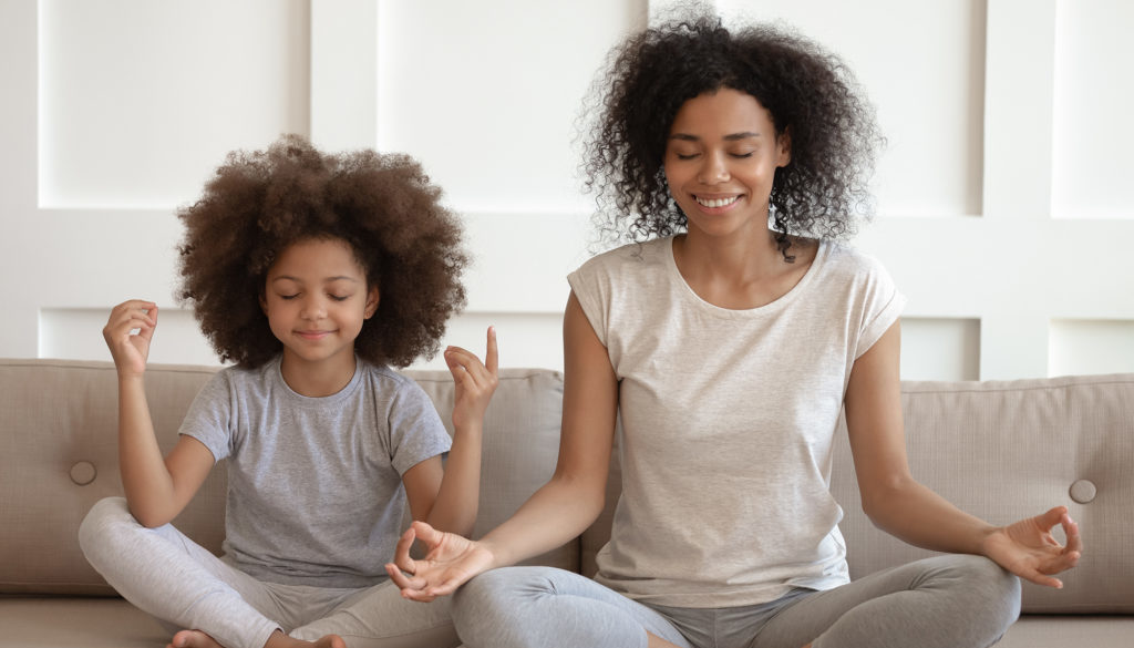 Black child and woman sit together on a sofa with their legs crossed and eyes closed, meditating.