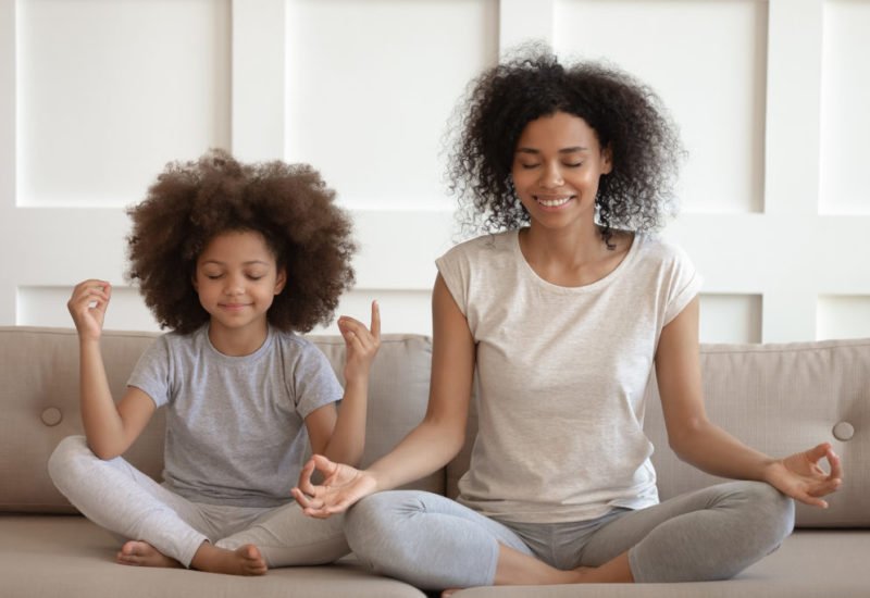 Black child and woman sit together on a sofa with their legs crossed and eyes closed, meditating.