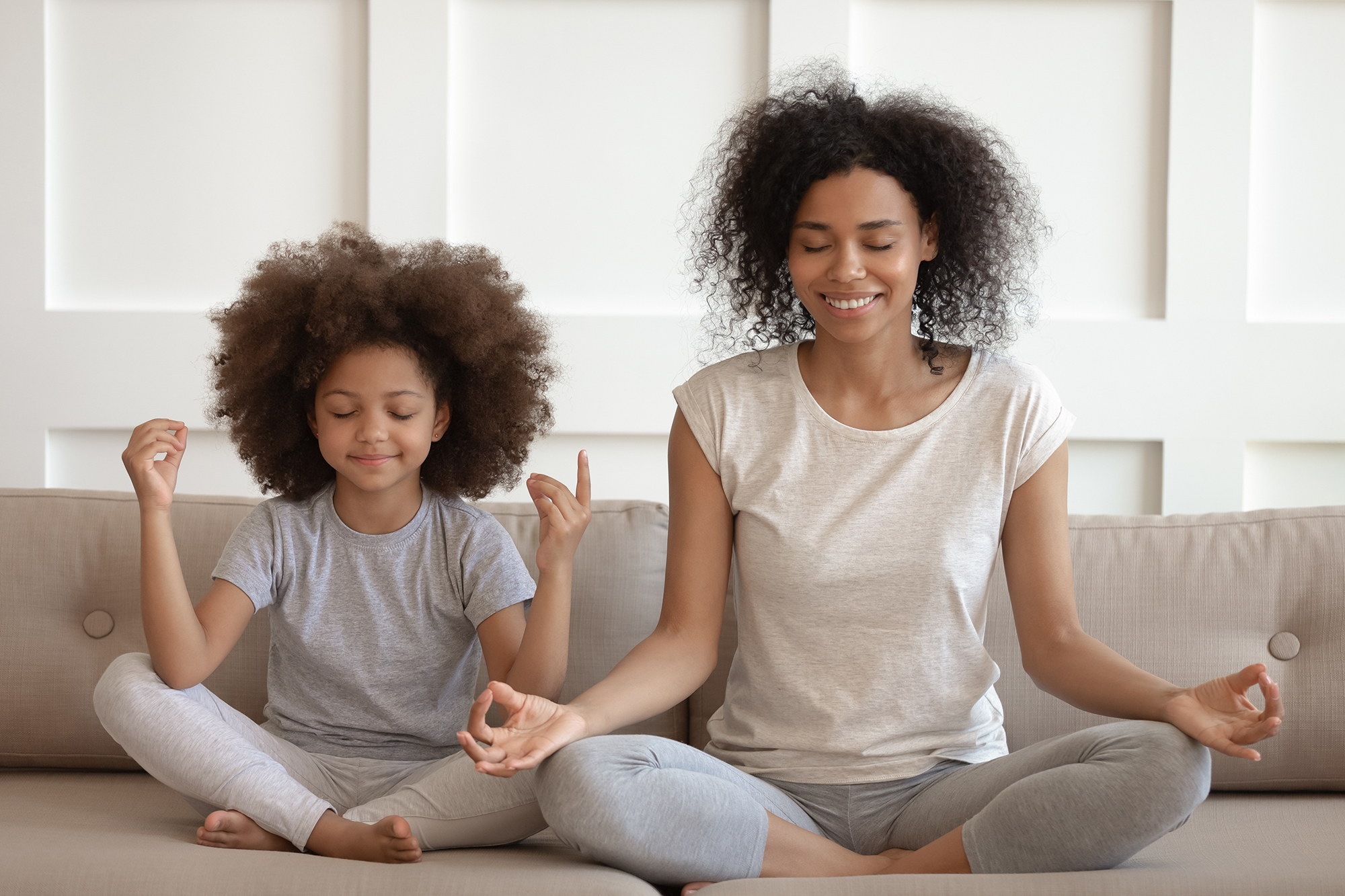 Black child and woman sit together on a sofa with their legs crossed and eyes closed, meditating.